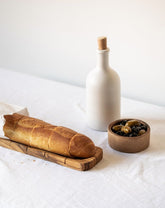 Olive wood bread slicing board with bread on top, accompanied by a snack bowl with contents and a cork-topped dispenser, set against a white background.